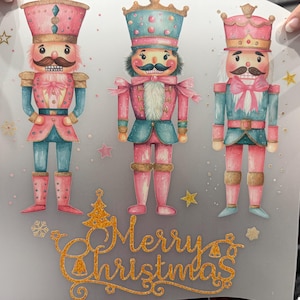 May include: A decorative print featuring three watercolour nutcracker figures in pink, blue, and gold. The figures have moustaches and are wearing hats and uniforms. The words "Merry Christmas" are written in gold script below.