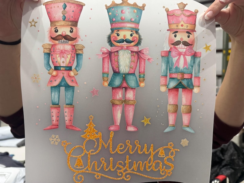 May include: A decorative print featuring three watercolour nutcracker figures in pink, blue, and gold. The figures have moustaches and are wearing hats and uniforms. The words "Merry Christmas" are written in gold script below.