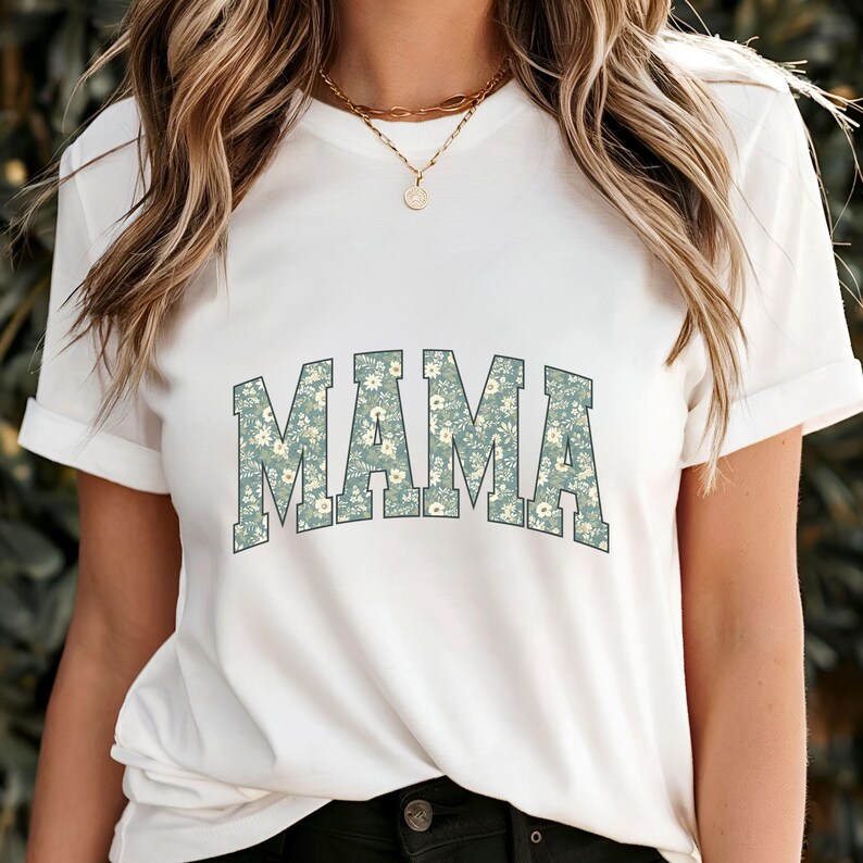 May include: White t-shirt with the word "MAMA" in a floral pattern across the chest. The letters are in a light blue-green color with a white and cream floral design. The shirt has short sleeves and a crew neck.