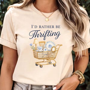 Op de afbeelding: Beige t-shirt met de tekst "I'D RATHER BE Thrifting" in marineblauwe letters. De afbeelding toont een gouden winkelwagen vol vintage items zoals een lamp, servies en een spiegel. Het ontwerp heeft een vintage uitstraling.