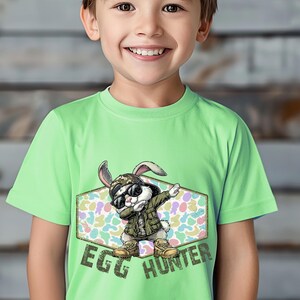 May include: A light green t-shirt featuring a cartoon rabbit wearing sunglasses, a military-style jacket, and boots. The rabbit is dabbing with the text "EGG HUNTER" below. The background is a pastel egg pattern.