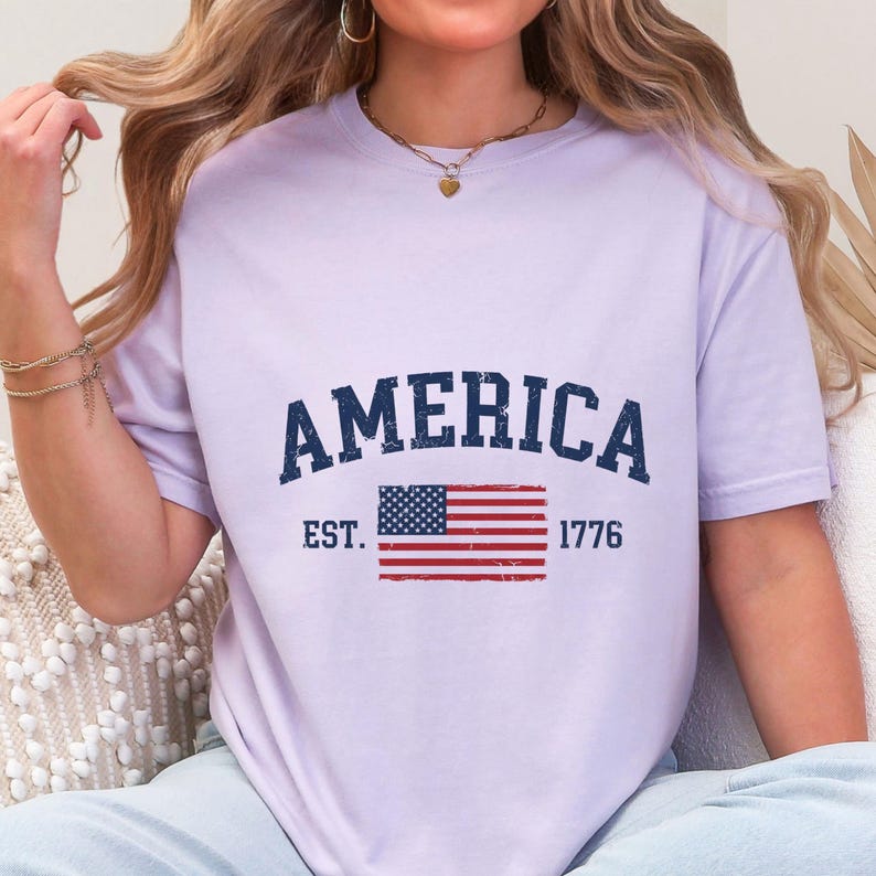 May include: A light purple short-sleeve t-shirt featuring the dark blue text AMERICA in a distressed collegiate font. Below the text is a graphic of the United States flag with the text EST. on the left and 1776 on the right in dark blue.
