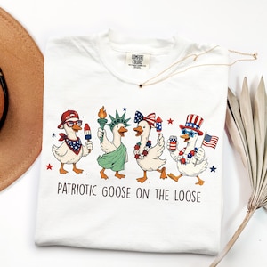 May include: White t-shirt with a patriotic goose design. The graphic features four cartoon geese dressed in red, white, and blue, holding items like a popsicle, the Statue of Liberty torch, and a can. Text reads "Patriotic Goose on the Loose."