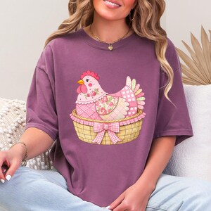 May include: A mauve t-shirt with a charming illustration of a patchwork hen in a woven basket with a pink bow. The hen has floral and polka dot patterns, and the basket has a gingham bow.