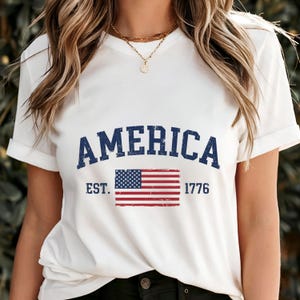 May include: A white short-sleeved t-shirt featuring the dark blue distressed text AMERICA arched above a graphic of the United States flag. The text EST. and 1776 are printed in dark blue on either side of the flag, which has a weathered texture.