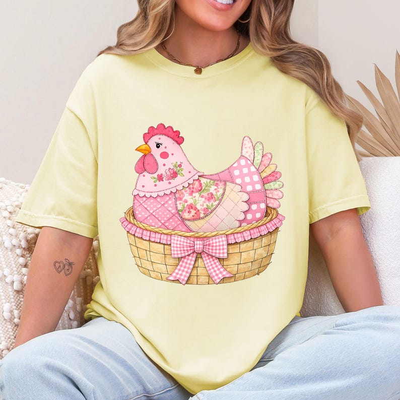 May include: A pale yellow t-shirt with a charming illustration of a pink patchwork hen in a woven basket, adorned with a pink gingham bow. The hen has a red comb and beak, and the basket has a ruffled edge.