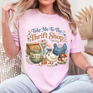 May include: Pink t-shirt with the phrase "Take Me To The Thrift Shop" and a graphic of a duck, chicken, bowls, bread box, and mushrooms. The design features floral and botanical elements, creating a vintage aesthetic.