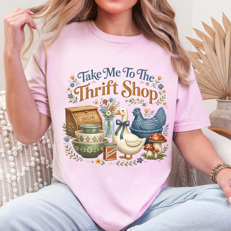 May include: Pink t-shirt with the phrase "Take Me To The Thrift Shop" and a graphic of a duck, chicken, bowls, bread box, and mushrooms. The design features floral and botanical elements, creating a vintage aesthetic.