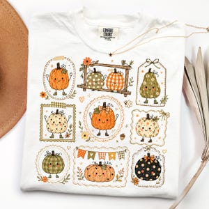 May include: A white t-shirt featuring a graphic print of various cartoon pumpkins with smiling faces, each enclosed in decorative frames or tags. The pumpkins display different patterns including polka dots, plaid, and solid orange, accented with small flowers, leaves, and a festive bunting banner.