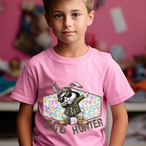 May include: Pink t-shirt featuring a cartoon rabbit wearing sunglasses, a military jacket, and boots, dabbing. The graphic is set within a hexagonal frame with a pastel egg pattern. The text "EGG HUNTER" is below the rabbit.