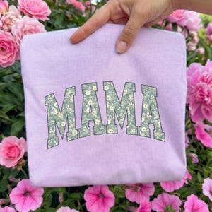 May include: A light purple sweatshirt with the word "MAMA" in large, floral-patterned letters across the front. The floral pattern features white and light green flowers on a green background. The sweatshirt is held up by a hand.