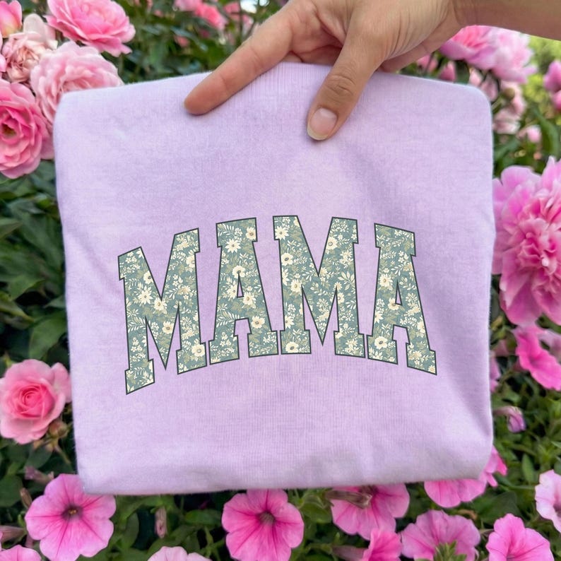 May include: A light purple sweatshirt with the word "MAMA" in large, floral-patterned letters across the front. The floral pattern features white and light green flowers on a green background. The sweatshirt is held up by a hand.
