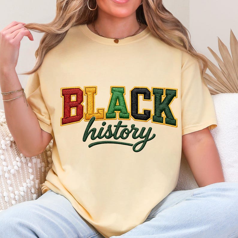 May include: A light yellow t-shirt with the words "BLACK history" in a colorful, embroidered design. The word "BLACK" is in red, yellow, green, and black, with a green embroidered "history" underneath.