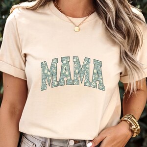 May include: A beige t-shirt with the word "MAMA" in large, stylized letters across the front. The letters are filled with a floral pattern in shades of blue and white. The shirt is being worn.