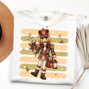 May include: A white t-shirt featuring a graphic of a nutcracker soldier in a red and green uniform, holding a candy cane and a lantern. The design includes horizontal brushstroke stripes in beige and green, gold stars, and white snowflakes.