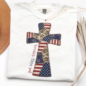 May include: White t-shirt featuring a cross design with American flag patchwork. The cross includes red, white, and blue stars and stripes, with the words "god bless america" in red script. The shirt has a crew neck and short sleeves.