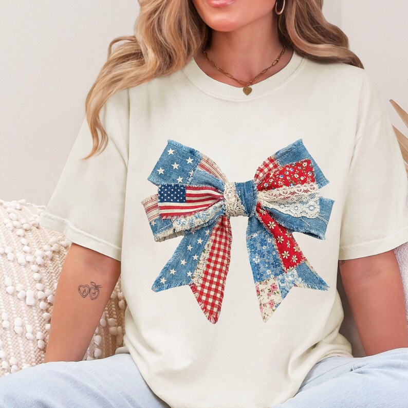 May include: Cream-colored t-shirt featuring a large bow design. The bow is crafted from various fabrics, including denim, red gingham, and floral patterns, with an American flag centerpiece. The bow is a patriotic and decorative element.