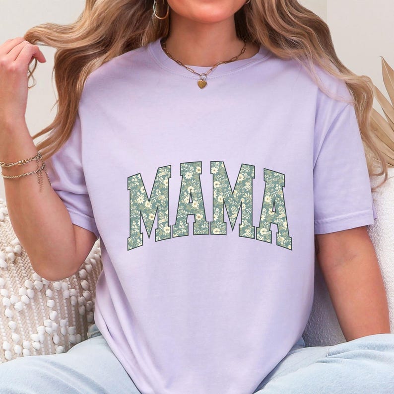 May include: A light purple t-shirt with the word "MAMA" in large, block letters. The letters are filled with a floral pattern in shades of green and white. The shirt has short sleeves and a relaxed fit.