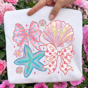 May include: White sweatshirt featuring a colorful beach-themed design. The graphic includes a blue starfish, pink bows with polka dots, a seashell with pearls, and floral accents. The design has a glittery gold outline.
