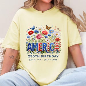 K&ouml;nnte beinhalten: Hellgelbes T-Shirt mit floralem Design und dem Wort "AMERICA" in Rot, Wei&szlig; und Blau. Das Shirt feiert den 250. Geburtstag mit den Daten 4. Juli 1776 - 4. Juli 2026.