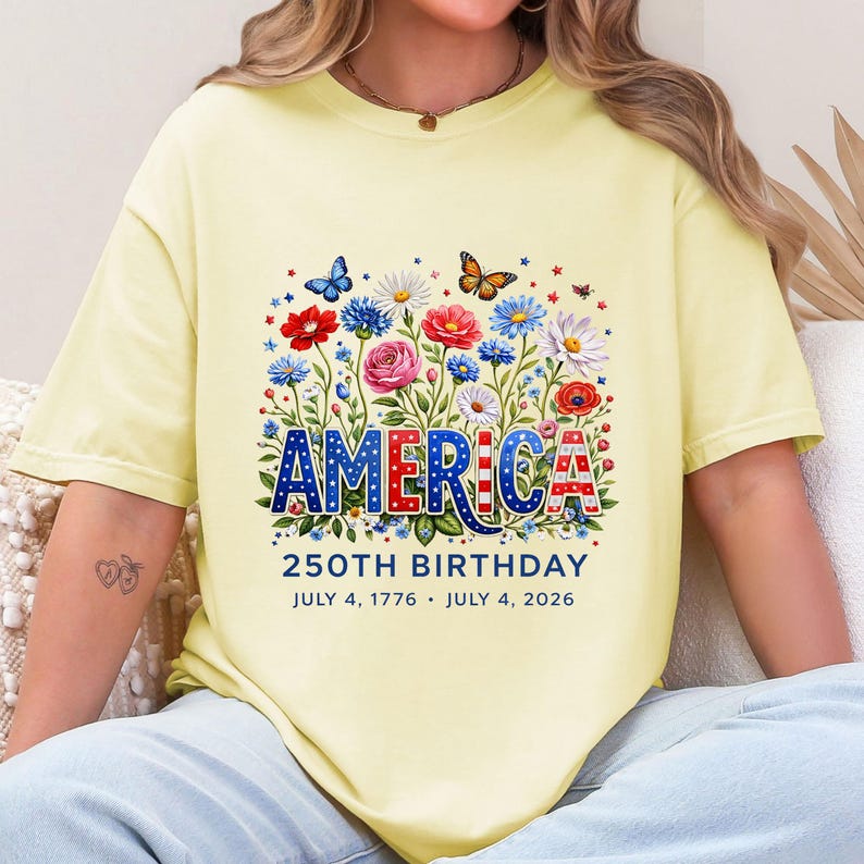 K&ouml;nnte beinhalten: Hellgelbes T-Shirt mit floralem Design und dem Wort "AMERICA" in Rot, Wei&szlig; und Blau. Das Shirt feiert den 250. Geburtstag mit den Daten 4. Juli 1776 - 4. Juli 2026.