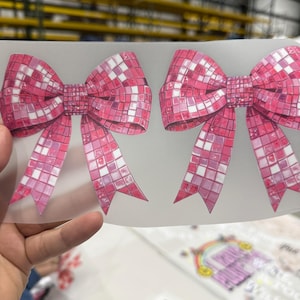 May include: Two pink bow-shaped stickers with a mosaic tile design. The bows have a gradient of pink hues, with white squares interspersed throughout. The ribbons of the bows are also pink and white. The stickers are on a clear backing.