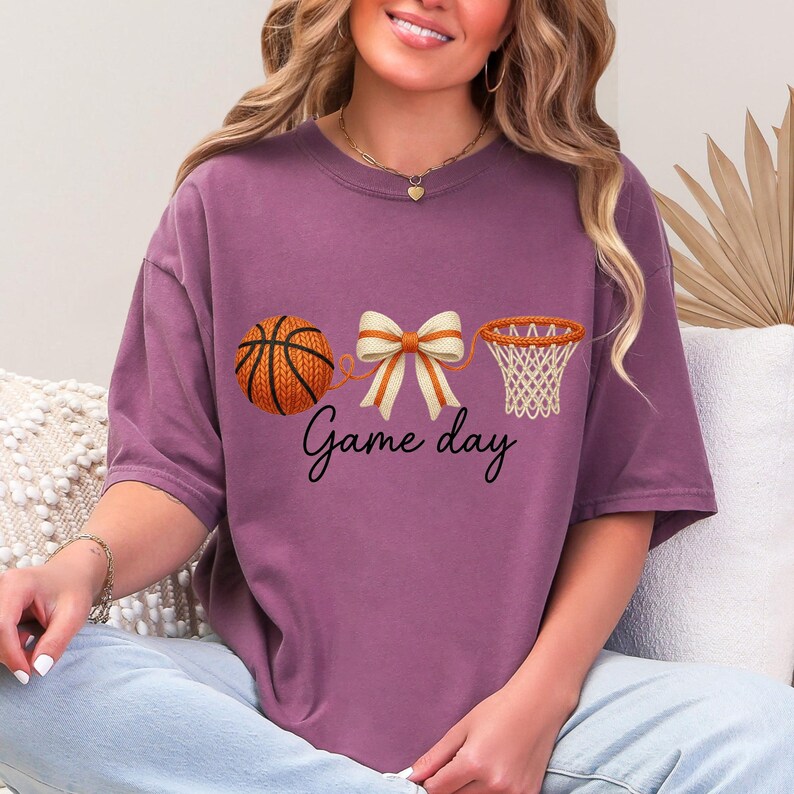 May include: A mauve-colored t-shirt featuring a basketball, a bow, and a basketball net graphic. The words "Game day" are written in a script font below the graphics. The basketball is orange and brown.