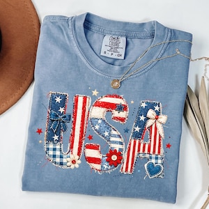 May include: A light blue Comfort Colors t-shirt with the word "USA" appliqued on the front. The letters are decorated with patriotic patterns, including stars, stripes, and bows. A gold necklace with a pendant is draped across the shirt.