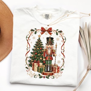 May include: A white t-shirt featuring a graphic print of a nutcracker standing next to a decorated Christmas tree and wrapped gifts. The design is framed by a red ribbon, holly leaves, and gold accents. The shirt has a Comfort Colors brand label.