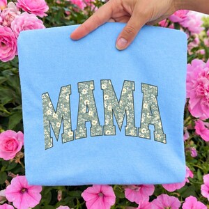 May include: A light blue sweatshirt with the word "MAMA" in a floral print design. The letters are in a collegiate style font. The sweatshirt is held up by a hand, surrounded by pink flowers.