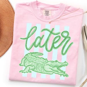 May include: Pink t-shirt with the word "later" in green script, over a green alligator illustration. Light blue vertical stripes are behind the text. The shirt has a crew neck and a Comfort Colors tag.