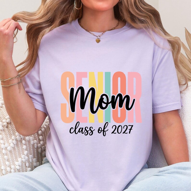 May include: A light purple short-sleeve t-shirt featuring the word SENIOR in large, vertical, multi-colored block letters behind the black cursive text Mom and the black printed text class of 2027 centered on the front of the garment.