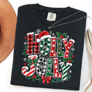 May include: A black t-shirt featuring the text HOLLY JOLLY in large, decorative letters. The letters are patterned with red and black plaid, green polka dots, white stars, red stripes, and white trees. A red Santa hat sits atop the H.