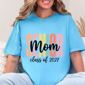 May include: A light blue short-sleeve t-shirt featuring the word SENIOR in large, vertical, multi-colored block letters behind the black cursive text Mom and the black printed text class of 2027 centered on the front of the garment.