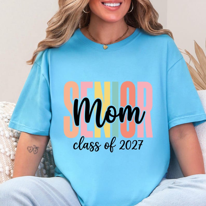 May include: A light blue short-sleeve t-shirt featuring the word SENIOR in large, vertical, multi-colored block letters behind the black cursive text Mom and the black printed text class of 2027 centered on the front of the garment.