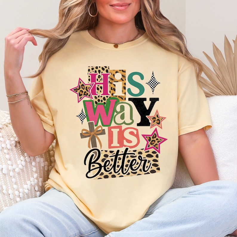 May include: A pale yellow t-shirt featuring the text "HIS WAY IS Better" in a vibrant, layered design. The design incorporates stars, a cross, and leopard print patterns. The shirt is a short-sleeved, casual style.