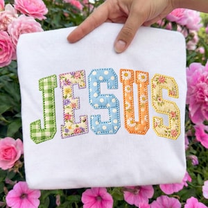 May include: White sweatshirt with the word "JESUS" embroidered in colorful, patterned letters. The "J" is green gingham, the "E" floral, the "S" blue polka dots, the "U" orange with daisy accents, and the final "S" floral.