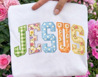 Puede incluir: Sudadera blanca con la palabra "JESUS" bordada en letras coloridas y estampadas. La "J" es de cuadros verdes, la "E" floral, la "S" de lunares azules, la "U" naranja con detalles de margaritas y la última "S" floral.