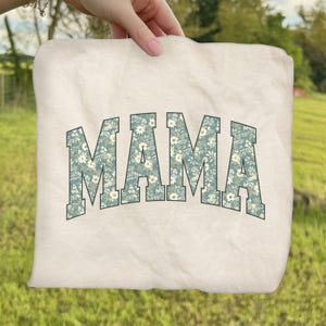 May include: Cream-colored tote bag with the word "MAMA" in large, block letters. The letters are filled with a floral pattern in shades of blue, green, and white. The bag is held up against a blurred green background.