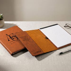 May include: A brown leather portfolio with a notepad, pen, and glasses. The portfolio is open, revealing a lined notepad. The cover is embossed with initials and the name "ALEX GARNER". The inside flap has text and the name "A. GARNER".