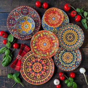 Puede incluir: Una colección de platos y cuencos de cerámica redondos con intrincados y coloridos patrones estilo mandala en tonos rojo, amarillo, azul y naranja. Una servilleta de cuadros rojos con un anillo dorado, tomates frescos y albahaca están dispuestos.