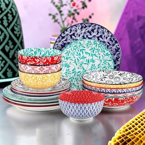 May include: A collection of ceramic dinnerware featuring various bowls and plates with colourful patterns. The set includes pieces with red, yellow, blue, and black floral, geometric, and leaf motifs, stacked and arranged in a decorative display.