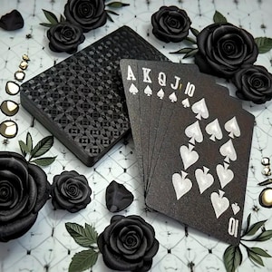 Puede incluir: Una baraja de cartas negras con símbolos de picas blancos y figuras, abanicada para mostrar una escalera real. La baraja y las cartas están rodeadas de rosas negras y elementos decorativos.