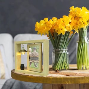 Puede incluir: Un marco de fotos de madera de color claro con una foto giratoria, una pequeña lámpara y un jarrón negro. El marco está sobre una mesa de madera con un ramo de narcisos amarillos en un tarro de cristal.