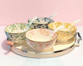 May include: A set of four ceramic bowls and matching spoons arranged on a round silver tray. Each bowl features a distinct floral pattern in green, blue, pink, or yellow with gold-toned rims. The spoons are white with floral accents.