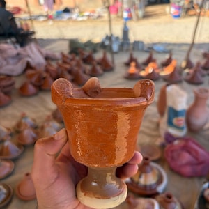 Puede incluir: Un cáliz de cerámica marrón hecho a mano con un borde ancho y una base de pedestal. El cáliz se sostiene en la mano de una persona, con otros artículos de cerámica similares visibles en el fondo, lo que sugiere un entorno de mercado.