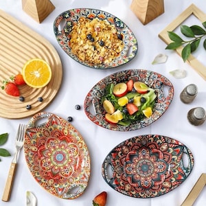 Puede incluir: Cuatro fuentes de servir ovaladas de cerámica con asas integradas, con intrincados y coloridos patrones de estilo mandala en tonos rojos, naranjas, azules y verdes. Dos fuentes contienen alimentos, una con cereales y arándanos, otra con ensalada de frutas.