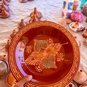 Puede incluir: Un plato de cerámica marrón rojizo con un diseño floral es sostenido por una mano. El plato tiene un borde decorativo naranja y blanco. Otros tagines y tapas de terracota son visibles en el fondo.