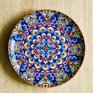 Puede incluir: Plato decorativo de cerámica con un diseño floral simétrico y vibrante. El plato presenta una rica paleta de colores azul, rojo, amarillo y naranja, creando un patrón visualmente impactante. El plato es redondo y adecuado para servir alimentos o como pieza decorativa.