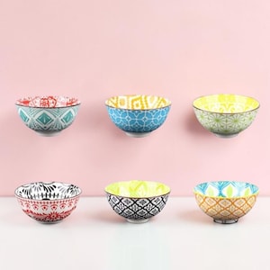 Puede incluir: Un conjunto de seis cuencos de cerámica dispuestos en dos filas de tres. Cada cuenco presenta un patrón colorido distinto, incluyendo diseños geométricos y florales en rojo, azul verdoso, azul, amarillo, verde, negro y naranja sobre base blanca.